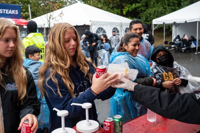 A white female with long dirty blonde hair, wearing navy blue Kean University sweatshirt that says Student Life in white letters, is being handed a Coca Cola, and basket of French fries that she ordered. The food tents are set up in the Harwood arena parking lot, and others who came to the event are gathered around the parking lot, getting food. 