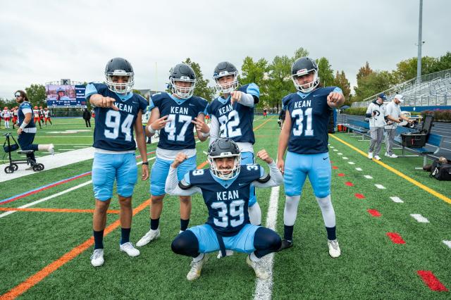 5 Kean football players are posing for a photo. They are all wearing Navy blue jerseys, and baby blue football pants, with cleats and helmets on. 4 players are standing, and 3 out of the 4 are pointing to the camera. The 4th player is throwing up both peace signs, and the 5th player is squatting in the center of the other 4, flexing both his biceps and smiling. They are standing on the football field, which has green turf grass.
