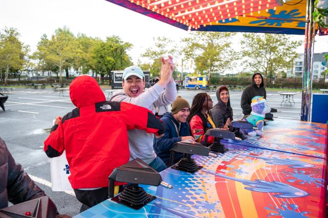 A diverse group of students wearing sweatshirts and hoodies are each sitting at their own chair in front of a water gun carnival game. The second student closest to the picture is standing up, clapping and celebrating (assuming he won the round). The student to his right (left in the picture) has his back to the camera, wearing a red north face jacket, congratulating the student who won. This event is set up in the Harwood parking lot at Kean.