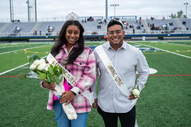 On the left stands the Homecoming Queen, a Black female with long black hair wearing a pink and white plaid jacket, a white homecoming sash with gold letters, and a silver tiara. She is holding a bouquet of white roses. On the right side of her stands the Homecoming King, a Latinx male with curly brown hair on the top, and a buzzcut on the sides, with a clean shaved black beard, wearing a light gray button up with silver glasses. He is wearing a white homecoming sash with gold letters, a silver King’s crown