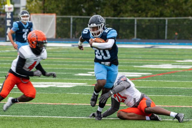 Kean football athlete, a Black male wearing a navy blue jersey with the number 20 in white, and baby blue football pants with black cleats and a navy blue helmet. He is holding the football with his right arm, running from two of the players of the opposing team. One opposing player seemed to try to tackle him, but missed. The other opposing player is on the left side of the Kean athlete in the photo. The Kean athlete is running on the football field, which is green turf grass.