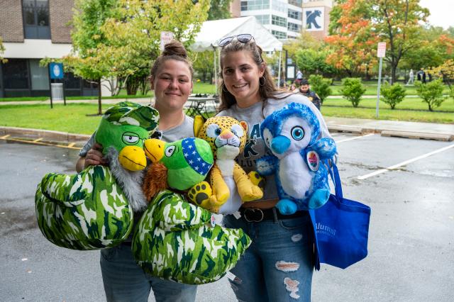 On the left, a white female with brown hair in a bun on the top of her head, wearing a gray short-sleeved shirt and jeans, holding 3 stuffed animal prizes, smiling at the camera. On the right, a white female with long brown hair, wearing a gray short-sleeve shirt with jeans, holding a blue colored bird stuffed animal, and blue Kean University bag. Both females are smiling for the photo in front of a parking lot that is surrounded by green trees. 