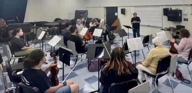 A conductor stands in the front of a rehearsal room filled with orchestral musicians.
