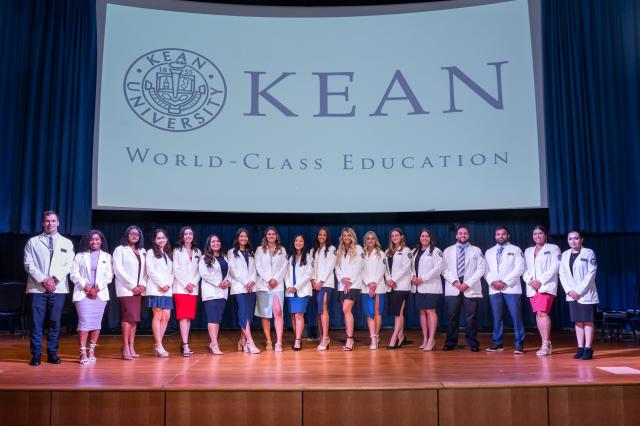 A diverse group of male and female Physician Assistants from Kean University. Females are wearing dresses and heals, while males are wearing suits and dress shoes. All students are wearing white lab coats. They are all standing side-by-side on the Kean University stage, with a white backdrop of the Kean logo written in navy blue letters. 