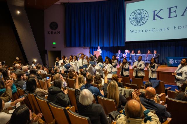  A diverse group of male and female Physician Assistants from Kean University. Females are wearing dresses and heals, while males are wearing suits and dress shoes. All students are wearing white lab coats standing in front of the stage, smiling and clapping, while looking into the crowd at their friends and family, as they clap and take pictures of the students. 