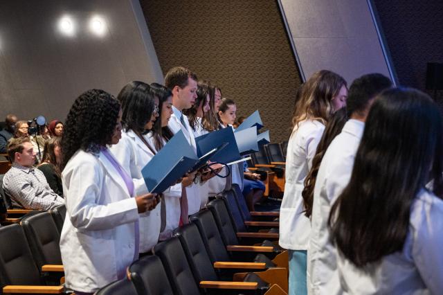 A picture of a diverse group of male and female Physician Assistants from Kean University. Females are wearing dresses and heels, while males are wearing suits and dress shoes, taken from the center of the aisle, looking to your left towards the seats of an auditorium. Students are holding navy-colored booklets, reading off of them. 