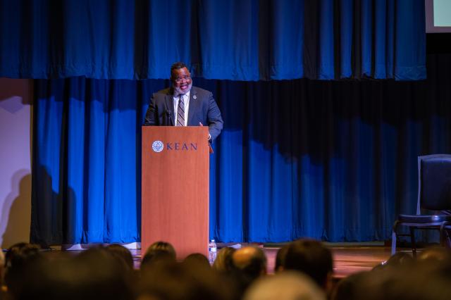 A wide-shot of President Lamont Repollet, a Black male with dark hair, and a gray and black beard and mustache wearing a dark navy suit and gold and navy tie that has a design on it, along with navy stripes and glasses. He is standing behind a podium with a microphone attached, speaking to the friends and family members who attended this event. 