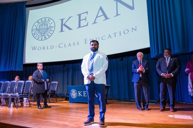 A wide-shot of an Indian male Physician Student with dark, long hair on the top, and a dark beard and mustache. He is wearing a whitecoat, a white collared shirt underneath with a blue and gold striped tie, and blue pants. He is also wearing brown dress shoes. To the right of him in the picture, is President Lamont Repollet, a Black male with dark hair, and a gray and black beard and mustache wearing a dark navy suit and gold and navy tie that has a design on it, along with navy stripes and glasses, and he 