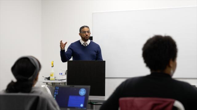 A wide-shot of Rafael Mata, regional director of the New Jersey SBDC at Kean, a Hispanic male with dark black and gray hair, and a dark black and gray beard and mustache, wearing a navy sweater, with a white button-up underneath, and a tan and brown striped tie with gray work pants and glasses. He is standing in front of a podium, gesturing to the individuals that attended this event.