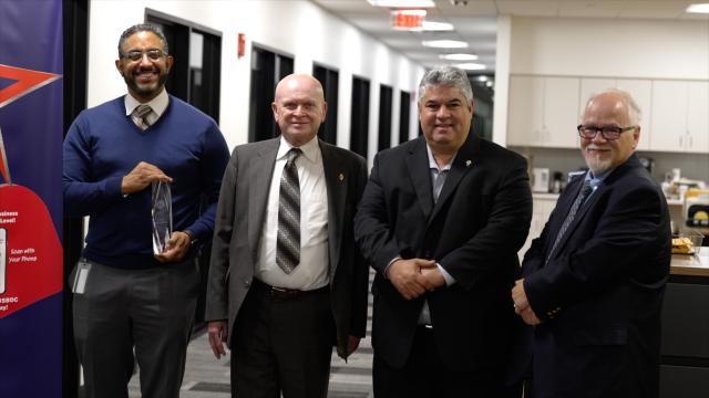  (L-R) Rafael Mata, regional director of the New Jersey SBDC at Kean, a Hispanic male with dark black and gray hair, and a dark black and gray beard and mustache, wearing a navy sweater, with a white button-up underneath, and a tan and brown striped tie with gray work pants and glasses. He is holding a see-through 12 inch award. Next to him is John Cronin, a state certification auditor, a bald, white male, wearing a dark gray suit, white button-up, and a gray, brown, and white striped tie. Next to John is L