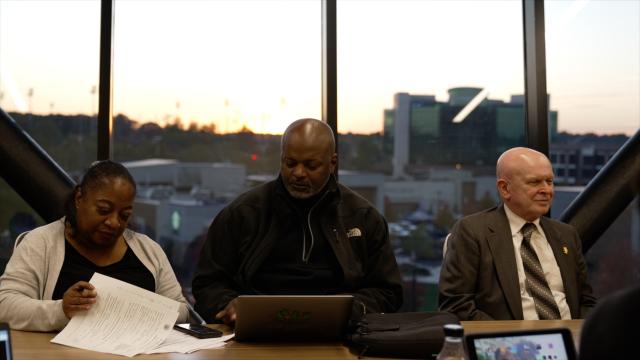 (L-R) a Black female with black hair pulled back, wearing a tan sweater and black shirt underneath is flipping through papers at the desk she is sitting at. Next to her is a bald, Black male wearing an all-black North Face jacket, and black shirt underneath, looking down at his computer. Next to him is John Cronin, a state certification auditor, a bald, white male, wearing a dark gray suit, white button-up, and a gray, brown, and white striped tie. All three of them are sitting behind a table, in front of a