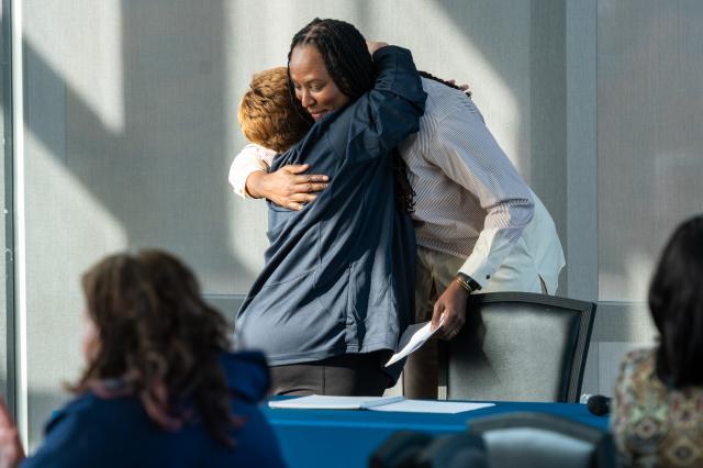 Chamique Holdsclaw, a Black female with long, black dreads, wearing a cream-colored button-up collared shirt, hugging a Kean Faculty member, a white female with short, brownish-red hair, wearing a navy blue long-sleeve jacket and black pants. This female’s back is towards the camera, but Chamique is facing towards the camera. They are both hugging behind a table with a blue tablecloth and a black chair behind it. 