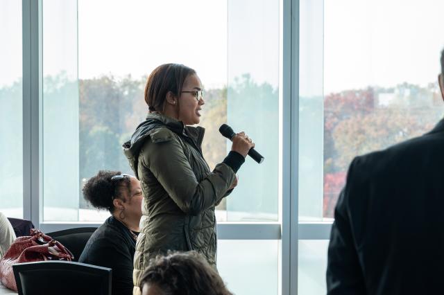 A Black female with brown hair that is pulled back, wearing glasses and a long, green jacket, is standing and holding a microphone with her right hand. Only her right profile is being shown, and she is talking into the microphone. She stands with the crowd, who are all seated, and there are glass windows in the background.