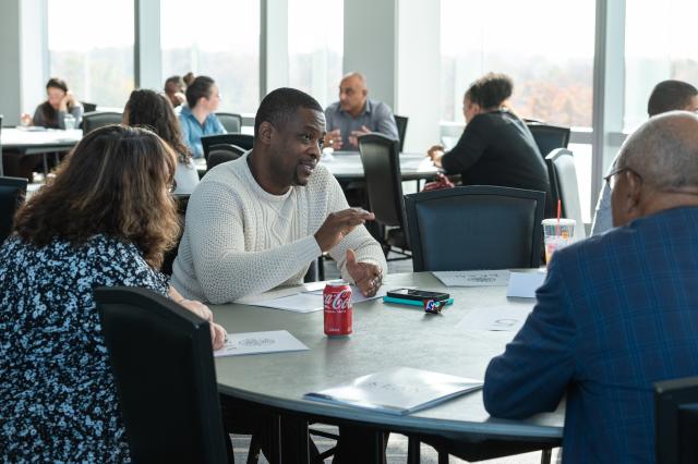 Pictured are 3 Kean faculty members sitting at a round table. The only face we can see is a Black male with black hair and a black beard and mustache, wearing a white-knit long-sleeve sweater, gesturing to the two other members at the table. On the left of the photo, a white female with short, brown wavy hair wearing a black and blue blouse is looking towards the Black male. To the right of the photo, sits a Black male with little gray hair, wearing a royal blue and navy blue striped blazer and black glasse