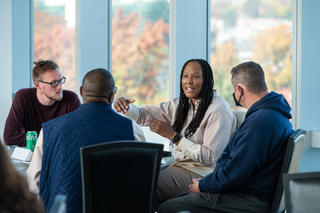 Chamique Holdsclaw, a Black female with long, black dreads, wearing a cream-colored button-up collared shirt, is gesturing to three other Kean University faculty members. They are all sitting in a half-circle at a glass table, sitting in black chairs. On the left, a white male with black glasses, light brown hair, and a light beard and mustache wearing a long-sleeve maroon shirt is looking towards Chamique, across from him sits a Black male with glasses, wearing a navy blue vest and cream colored long-sleev