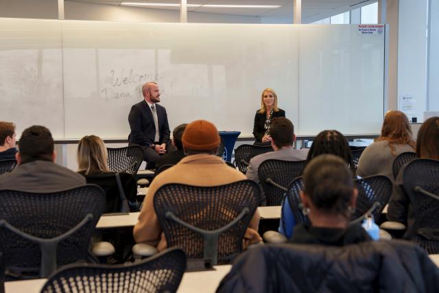 A wide-shot of Dana Bash, a white female with short, flowy blonde hair, wearing an all-black suit, and printed blouse underneath, sitting on a high-chair across from Acting Director of Career Curriculum and Academic Partnerships Jeremiah Sullivan, a white male with short brown hair, wearing an all-black suit, sitting on a high-chair as well with a round, high table in between them both. Pictured are the backs of Kean University students who are sitting at lecture tables, listening to Dana Bash.