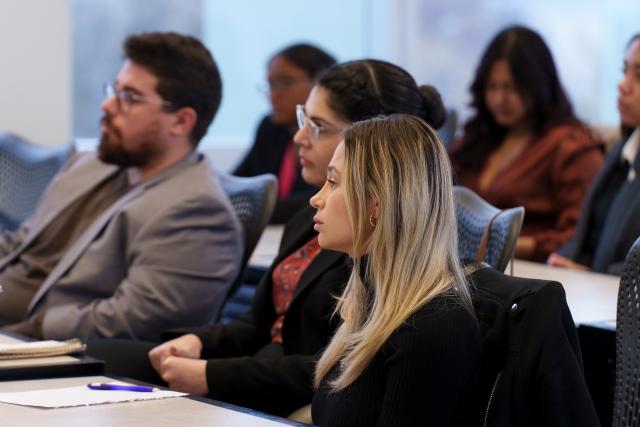 A side-profile shot of Kean University students, one male and two females sitting at a gray desk. Closest to the camera is a white female with long, blonde hair, wearing a black blazer. In between her and the male is a Latinx female with dark hair in a bun, wearing clear glasses, and a black blazer. On the farside is a white male with dark, short hair, and a dark mustache and beard wearing glasses, and a gray blazer.