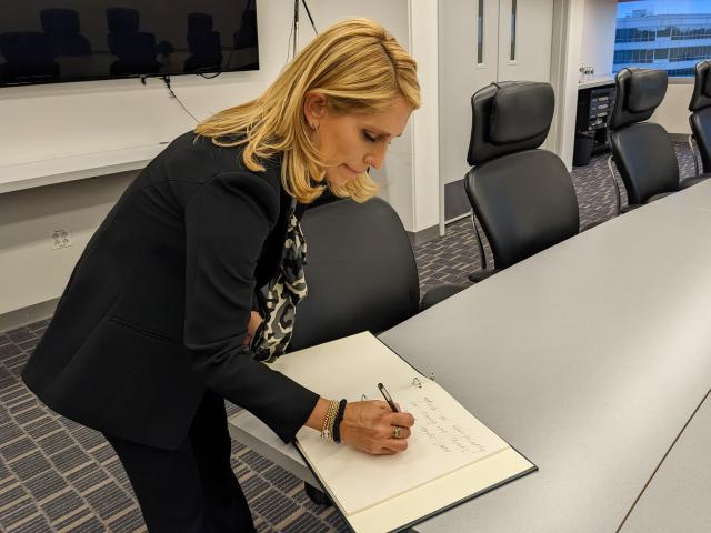 Dana Bash, a white female with short, flowy blonde hair, wearing an all-black suit, and printed blouse underneath, signing a book that previous Distinguished Lecturers have signed. She is standing, writing with her right hand.