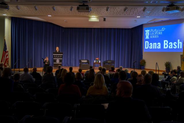A wide shot of Dana Bash, a white female with short, flowy blonde hair, wearing an all-black suit, and printed blouse underneath, is standing on stage, behind a black podium, speaking to the crowd of Kean faculty, staff and students. A long, navy current is behind her. A screen to the right of the shot is displaying Dana Bash’s name on a blue screen. 