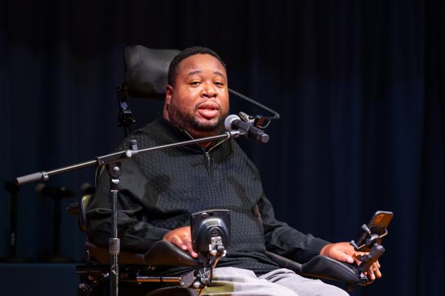  Close-up of Eric LeGrand, a Black male with short black hair, and a black beard and mustache, wearing a black long-sleeve quarter-zip and gray pants, sitting in his wheelchair speaking into a microphone to the audience.