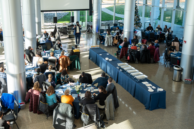 An overhead shot of a conference space with glass walls. A speaker is at the microphone, and round tables are filled with participants.