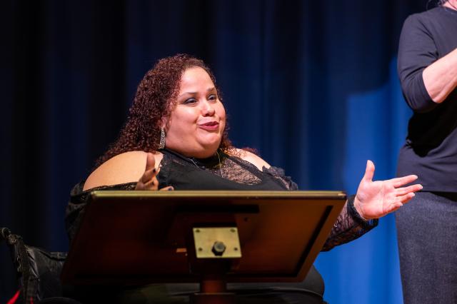 Close-up of Millie Gonzalez, a Latinx female with reddish brown curly hair that touches her shoulders, wearing a long-sleeve black and lace dress that has slits in it that shows her shoulders. She is wearing long, silver earrings and a silver necklace. She is in her black wheelchair, behind a podium, speaking to the audience. She is on stage, and blue curtain is behind her.