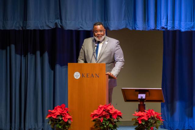 A close-up of President Lamont Repollet, a Black male with curly short black hair, and a gray beard and mustache wearing glasses and a gray suit and navy blue tie, standing behind a podium with a microphone attached, speaking to members who attended this event.