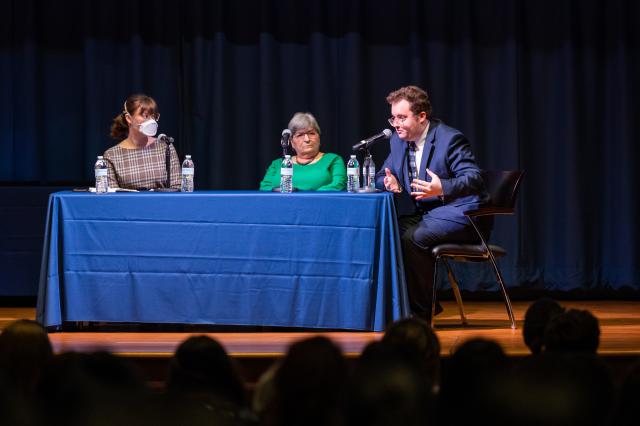  (l-r) a white woman with brown hair in a ponytail, wearing glasses and a brown and white plaid long-sleeve dress, she is also wearing a mask. Next to her is an elderly white woman with short, gray hair wearing a long-sleeve green dress, glasses, and a necklace. Next to her is a white male with curly brown hair and glasses, wearing a navy suit and white undershirt and blue tie. He is speaking into a microphone. Everyone has a microphone in front of them and they are sitting behind a table.