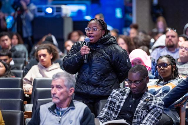  A Black female with black dreads pulled back, wearing clear glasses, and a black puffy coat, holding a microphone and speaking into it. She is standing in the center of the diverse audience.