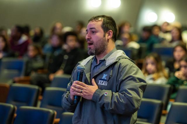 A white male with short brown hair, and a brown and gray beard and mustache, wearing a gray long-sleeve rain jacket that has the kean logo on the top left of his chest, holding and speaking into a microphone. He is standing in the crowd. 