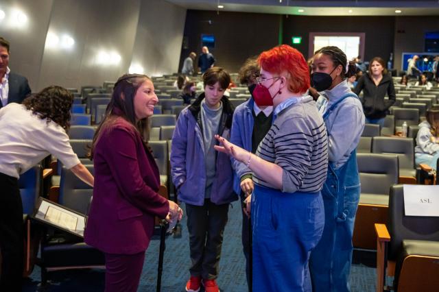 Isabel Mavrides-Calderón, a Latinx female with brown hair, wearing a maroon suit, stands with students who are asking her questions.