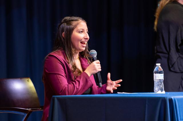 A Latinx female with long, brown hair that is half pulled back, wearing a pink blazer and speaking into a microphone that she is holding. She is sitting down in front of a table, looking towards the audience. She is on the Kean stage, with a blue curtain in the background.