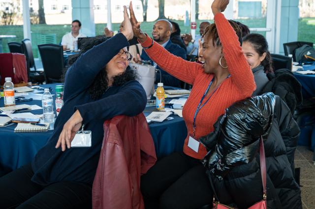 (L-R) a Latinx female with black, curly hair, wearing a navy blue long-sleeve and black glasses, sitting on a chair with a maroon jacket on the back. She is high fiving a Black female woman to the right of the photo, who has short brown dreads, wearing an orange long-sleeve shirt with silver hoops. They are sitting around a round table.