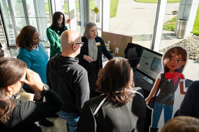 A shot of a diverse group of (5) females and (1) male, all gathered together in a circle, looking at a computer screen. There is a cardboard cut-out of a Black female cartoon dressed in casual clothes to the right of the computer. 