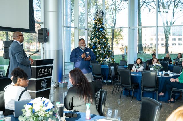 A wide-shot of President Lamont Repollet, a Black male with curly short black hair, and a gray beard and mustache wearing glasses and a navy blue quarter zip with gray pants. He is holding and speaking into a microphone out to the audience in the crowd. There is a Christmas tree decorated to his right with blue, white, and silver ornaments hanging and white lights.