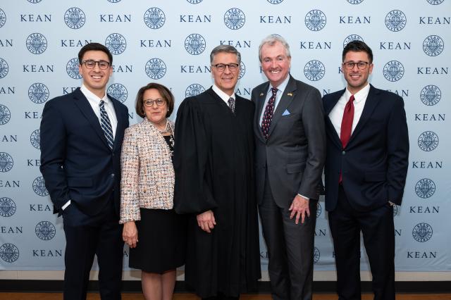 Governor Phil Murphy, a white male with gray hair, wearing a gray suit and light blue button-up and navy blue tie with Santa Claus on them, standing with (3) white males, and (1) white female, all wearing business professional clothing, standing side-by-side, smiling.