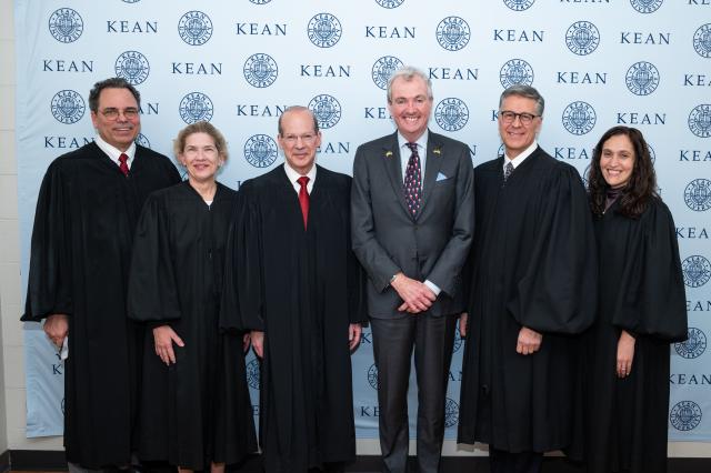 Governor Phil Murphy,  a white male with gray hair, wearing a gray suit and light blue button-up and navy blue tie with Santa Claus on them, standing in the middle of (2) white females, and (3) white males, all wearing long, black gowns.