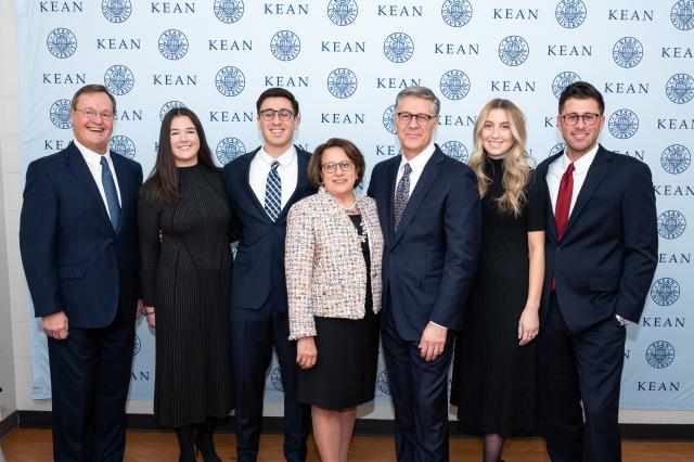 A group of (3) white females, and (4) white males, all dressed in business professional clothing. They are all standing together side-by-side, smiling.