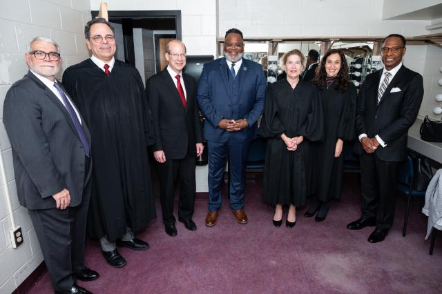 President Lamont Repollet, (L-R) President Lamont Repollet, a Black male with dark black hair and gray and black facial hair,  wearing glasses, a navy suit and white button-up shirt underneath and a blue and black tie, standing in the middle of (3) white males, (1) Black male, and (2) white women, some wearing business professional clothing, and others wearing long, black gowns.
