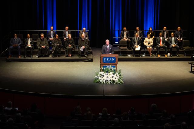 a wide-shot of the Kean University stage, and in the center behind a brown podium is a white male wearing an all-black suit. Behind him are a diverse group of men and women sitting on bleachers in black gowns. 