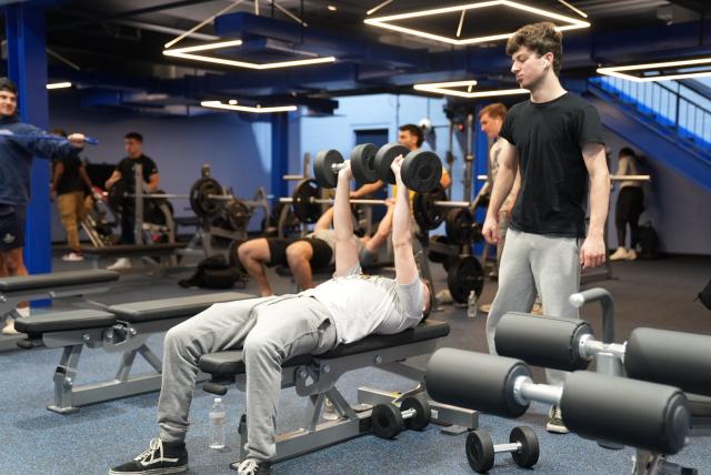 A wide-shot of a gym and a white male is laying on a black bench wearing a white t-shirt and gray athletic pants, benching two black dumbbells while his friend, a white male wearing a black t-shirt and gray athletic pants with brown hair is standing over him