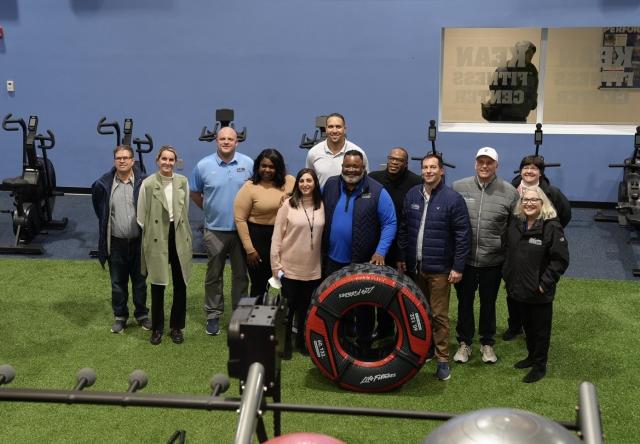 A birds-eye-view shot of Dr. Repollet,  a Black male with dark hair, and gray facial hair wearing navy pants, a blue long-sleeve, navy vest and athletic sneakers and glasses, standing with a diverse group of Kean employees wearing business attire, and business casual attire. Dr. Repollet is holding a black exercise tire in front of him. 