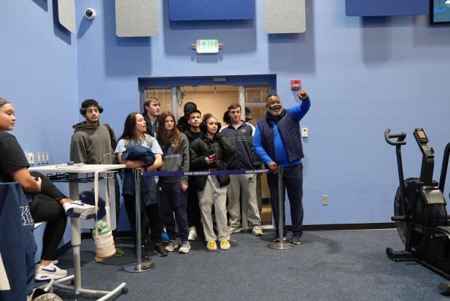  A diverse group of kean students standing inside the entrance to the new fitness center with President Dr. Repollet, a Black male with dark hair, and gray facial hair wearing navy pants, a blue long-sleeve, navy vest and athletic sneakers and glasses, waving his left arm.