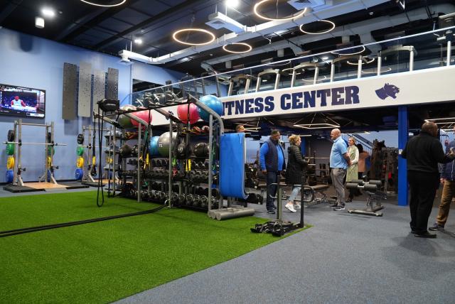 A wide-shot of Kean's brand new fitness center that includes green indoor turf, exercise balls, dumbbells, exercise bands in the center of the photo, and squat racks with weights to the left of the photo. Above says "fitness center" in navy blue, and above that is an upstairs with treadmills.