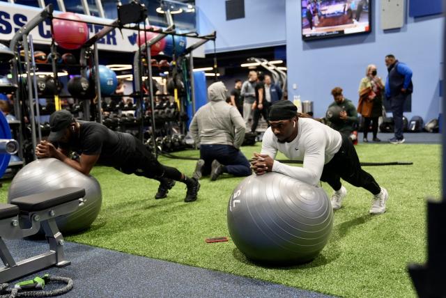 A wide-shot of two Black males (on the left) wearing a black t-shirt and black pants and a black hat, (on the right) wearing a white long sleeve and black pants, both doing a plank exercise on gray exercise balls, on a green indoor turf field
