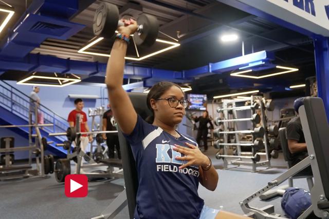 A young woman in a Kean t-shirt sits in a gym, lifting a small barbell over her head.