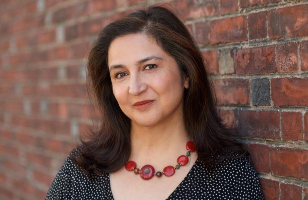 Rajika Bhandari, Ph.D., wearing a red necklace with black and white dress, standing in front of a brick wall. 