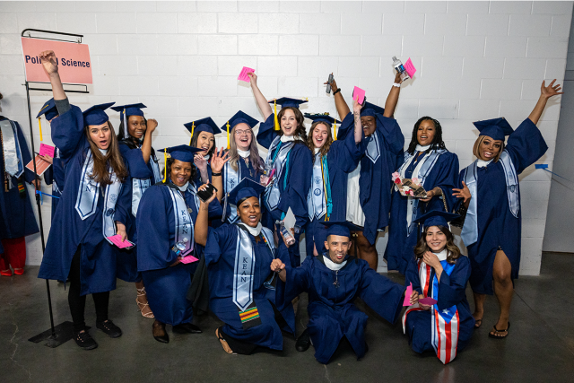 A group of Kean graduates, in blue caps and gowns, cheers with arms raised.