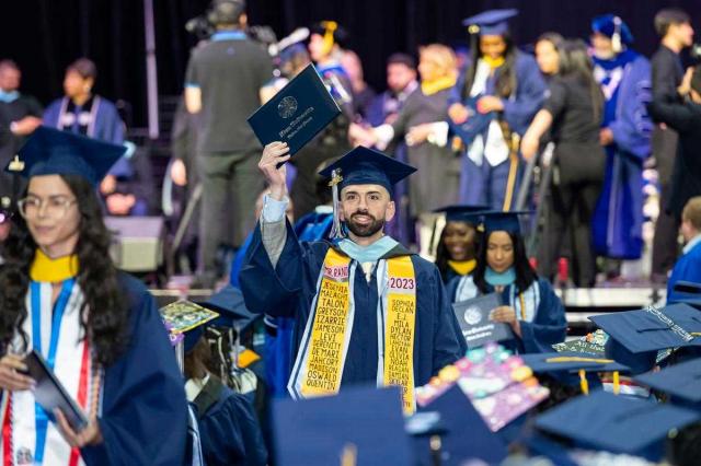A young man in a blue cap and gown raises his diploma certificate after walking off stage.