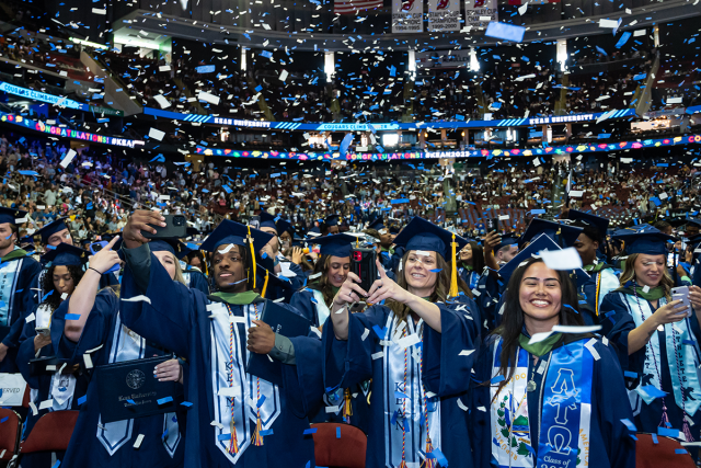 Kean graduates in blue caps and gowns celebrate as confetti blows through Prudential Center.